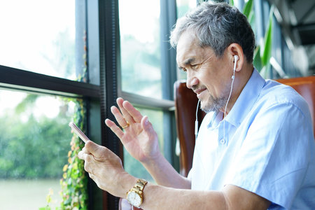 Asian senior man sitting among nature atmosphere, talking and waving to someone by online video call on mobile smartphoneの写真素材