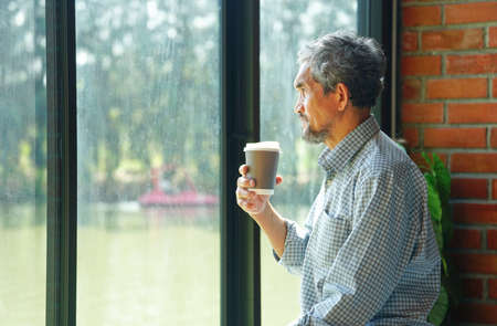 senior businessman is thoughtful and holding coffee cup in hand beside glasses window in nature atmosphereの写真素材
