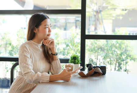 portrait young attractive asian woman traveller relaxing in the coffee shop in the morning sunshine  with smartphone in hand and camera on tableの写真素材