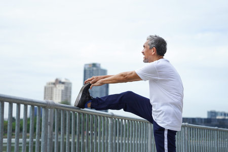 happy senior man with grey hair stretching legs in the city park, an old man warming up before...