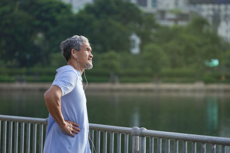 asian senior man with grey hair and beard wearing headphones for listening music is warming up in the city park before workout, concept elderly people lifestyle, healthcare, happiness, well beingの写真素材