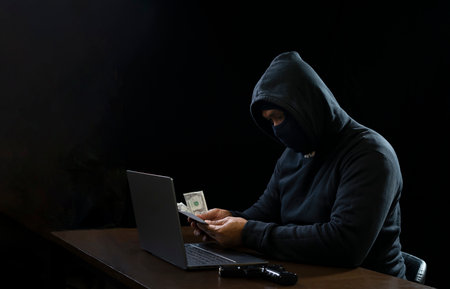cyber crime concept, robber in black hoodie holding money in hands and laptop with a gun put on tableの写真素材