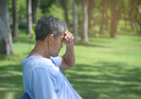 senior man hand holding his head, older adult male look headache sitting under the shade of trees in the park after workout, risk of overexercising in elderly peopleの写真素材
