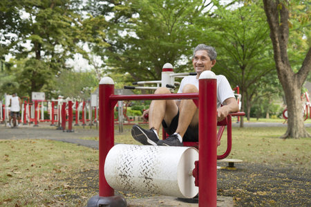happy senior man with grey hair doing legs muscles exercise on outdoor fitness equipment in the city...