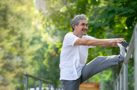 happy senior man with grey hair stretching his legs in the park, adult older warming up before exercise in the morning sunshine. concept for elderly pensioner people lifestyle, healthcare, wellbeingの写真素材