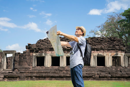 senior man with grey hair,carry backpack and having camera see a map of the ancient site while visit Phimai Historical Park,Thailand, concept eco-tourism, archaeological site, archeology,education,researchの写真素材