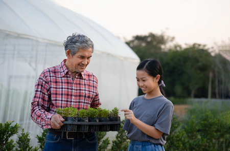 asian girl is interested in ornamental plants on nursery pot in senior man hands , grandfather is teaching granddaughter to know the trees that he planted at greenhouseの写真素材