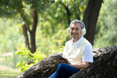 older adult male with grey hair wearing earphones to listen music hands holding tablet, senior man...