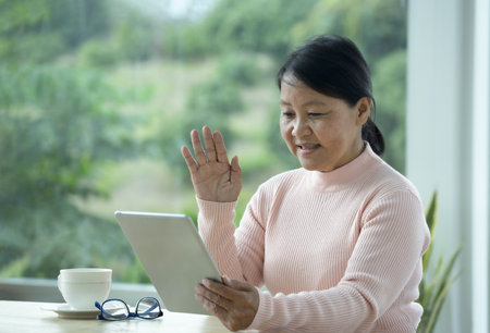 happy mature woman sitting by glass window, waving hand to greeting family by tablet online internet.concept internet, cell phones, family communication with loved ones both near and farの写真素材