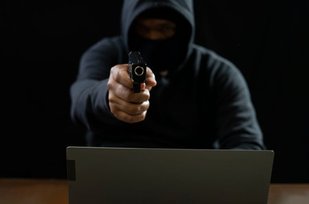 hacker in black hoodie with mask cover face poining a gun to camera, there is laptop on...