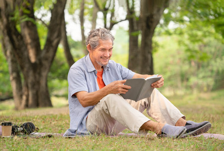 senior man with grey hair holding a book, smiling, sitting near a big tree in forest park. concept of elderly people lifestyle,hobbies,relaxing,health benefits in nature,wellbeingの写真素材