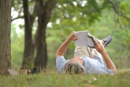old man with grey hair lying on grasses,smiling and reading a book in the park, concept of elderly lifestyle, hobbies, resting, relaxing, elder health benefits in nature,wellbeingの写真素材