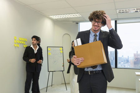 concept get fired, lose job, get stressed out.a man employee being fired holding a box of belongings leaving office, background colleagues are having a meeting.の写真素材