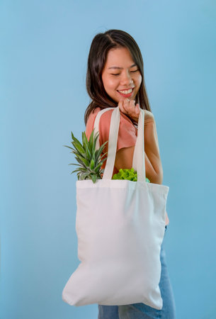 happy young asian housewife carrying reusable cloth bag of fruits and vegetables isolated on blue background,concept for campaign of using reusable bag, reduce waste,stop global warming,save the worldの写真素材