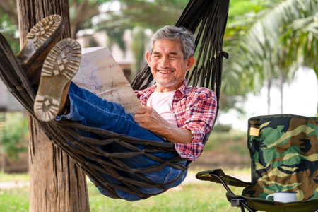 happy senior male with grey hair and beard wears checkered shirt relax at resort garden on summertime,mature man laying on rope hammock,smile and look at a paper map,concept of travel,camping,restingの写真素材