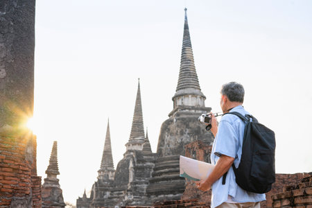 asian senior tourist male visit historic city,old man having a map,backpack,camera,is taking photo at Ayutthaya historical temple in evening sun,elderly people lifestyle,travel in archaeological siteの写真素材