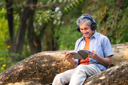 portrait happy senior man sitting on tree trunk in summer park,listening music and reading a book,peaceful old mature male relax outside in nature,concept of elderly people lifestyle,hobby,relaxationの写真素材