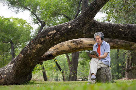 portrait happy senior man sitting on big log in summer park,listening music and reading a book,peaceful old mature male relaxing outside in nature,concept of elderly people lifestyle,hobby,relaxationの写真素材