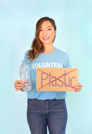 female volunteer in blue T-shirt,hand holds used plastic bottle,other hand holds cardboard sign no plastic,on blue background,concept campaign to recycle used empty plastic bottles,stop global warmingの写真素材