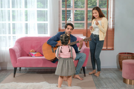 father sitting on sofa playing guitar,a little daughter looking at him while mother standing,sing a song,family spend time together in the living room,concept family,relationship,the love of a familyの写真素材