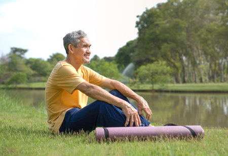 happy senior man in sportswear smiling and sitting in nature,older adult male with yoga mat resting by the pond in the park after yoga,concept of elderly people lifestyle,health care,relaxing,wellnessの写真素材