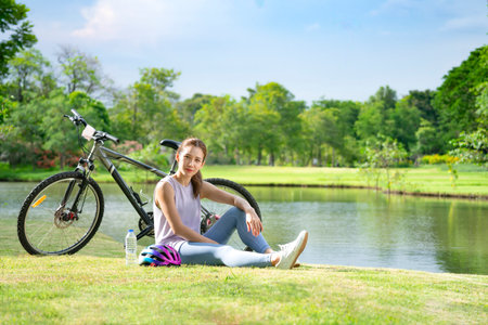 portrait attractive asian woman in sportswear sitting on grass in the park with a bicycle parked beside her,young sporty female resting by the pond in the morning sunshineの写真素材