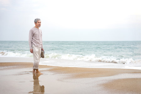 asian senior man barefoot,holding shoes walking on sand beach in the morning,healthy elderly male relaxing in nature,wellness,wellbeingの写真素材
