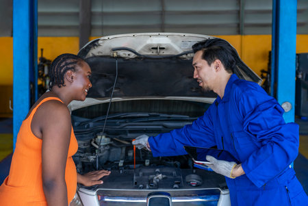 young asian auto mechanic male in workwear holding tablet computer,open a car hood explaining diagnosis of a car to african pregnancy female customer after finished examiningの写真素材