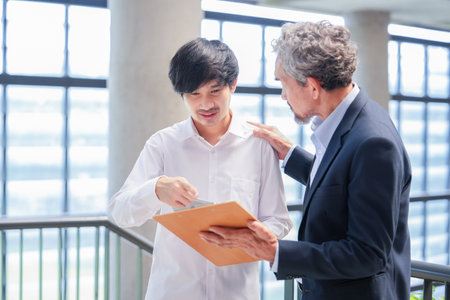 A young asian man standing and conversation with advisor teacher in a building,professor giving advice and university student with clipboard, support and touching his shoulderの写真素材