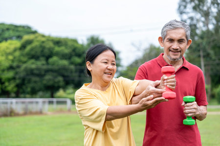 asian senior married couple doing exercise in the nature park together,elderly man support a wife to workout with dumbbellの写真素材