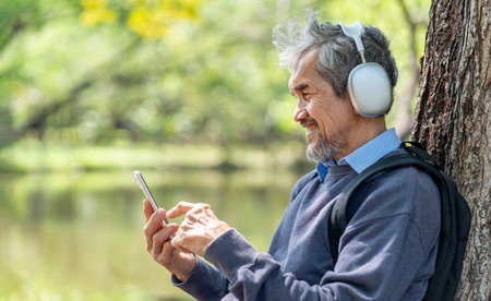 portrait happy asian senior man with grey hair and beard wears headset holding smartphone to listening music,sitting on bench in the parkの写真素材