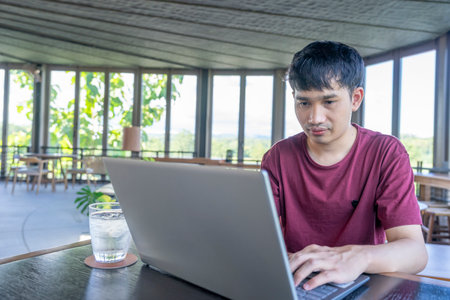 a young asian man in casual clothes remote working on laptop online internet at resort cafe,concept of travel,technology,working,digital nomadの写真素材