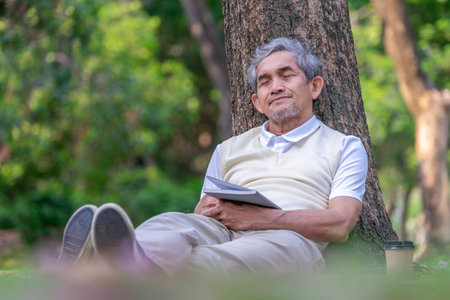 A tranquil senior man sleeping against a tree while reading a book in the park,の写真素材