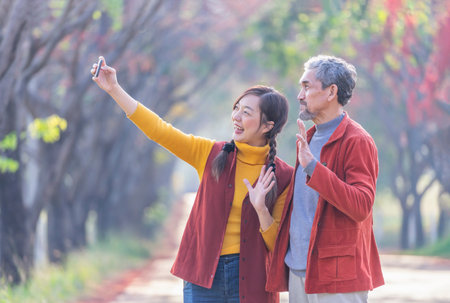 family lifestyle in autumn travel destination,young adult daughter and senior father smiling and taking a selfie together at the the park in fall seasonの写真素材