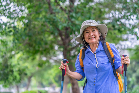 portrait cheerful senior woman wears hat,with backpack and trekking poles standing in nature,elderly asian female happy in hiking tripの写真素材