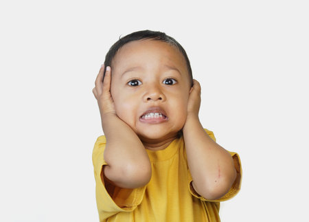 Portrait of a cute three year old Asian little boy with his hands on his head showing an expression of noise due to loud noise and fear, isolated on white backgroundの写真素材