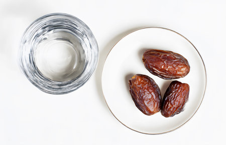 top of view Three dried dates on a white plate and a glass of water for takjil or appetizers when breaking the fast during the month of Ramadan, isolated on a white backgroundの素材