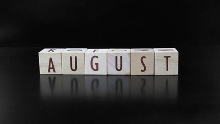 The word AUGUST is made of letters formed from wooden blocks, isolated on a black background.