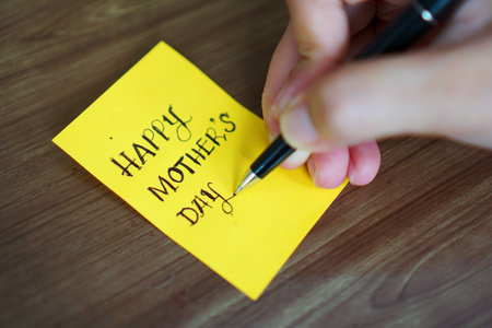 A woman is writing Happy Mother's Day to give to her mother on an orange sticky note using a black ballpoint pen on a brown table.の写真素材