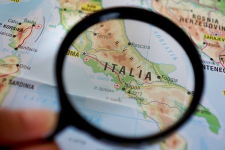 A woman's hand is holding a magnifying glass looking at a map of Italy, Europe on a geographical mapの写真素材