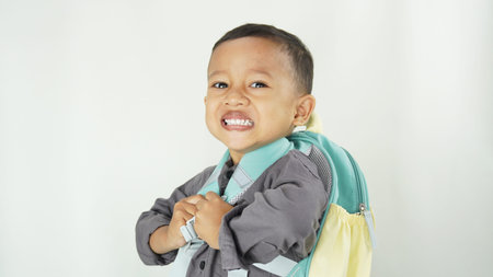 A cheerful Asian Muslim preschool-aged boy with his school bag and backpack wearing a gray...