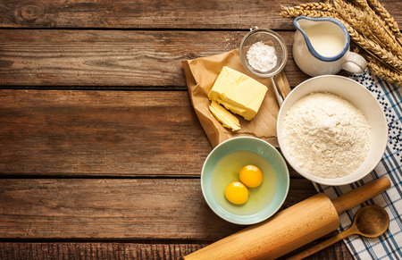 Baking cake in rural kitchen - dough recipe ingredients (eggs, flour, milk, butter, sugar) and rolling pin on vintage wood table from above.