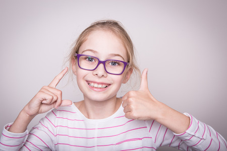 Happy smiling eight years old pretty blond caucasian child girl in violet glasses showing thumbs up gesture. Layout with free copy space.の写真素材