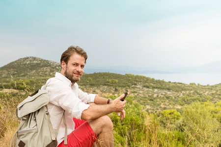 Happy smiling forty years old caucasian tourist man taking a break while mountain trip. Hills and sea as background. Summer vacation (holiday), traveling and adventure.の写真素材