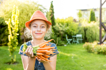 Happy smiling eight years old pretty blond caucasian child girl holding bunch of young carrots outdoor in the garden. Healthy lifestyle and gardening concept. Holiday - sunny summer day.の写真素材