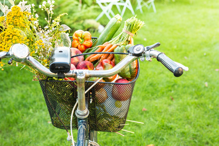 Bicycle basket filled with fresh vegetables and romantic wild-flower bunch. Gardening or healthy countryside lifestyle concept. Outdoor garden scenery as background. Sunny summer weekend mood.の写真素材