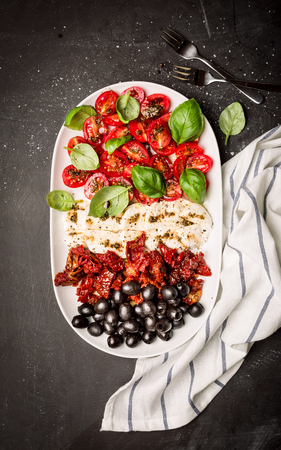 Italian appetizers - mozzarella cheese, sun dried and fresh cherry tomatoes, olives and basil on white plate. Black chalkboard as background. Flat lay composition (from above, top view).の写真素材