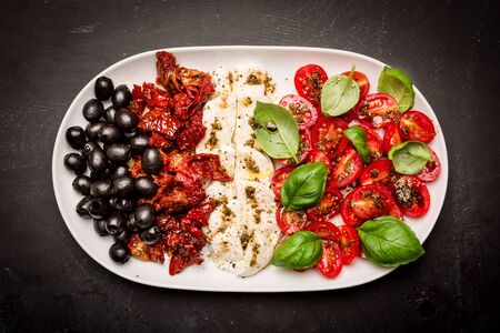 Italian appetizers - mozzarella cheese, sun dried and fresh cherry tomatoes, olives and basil on white plate. Black chalkboard as background. Flat lay composition (from above, top view).の写真素材