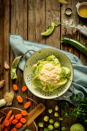 Cooking - fresh wet raw cauliflower in a strainer (colander) with other vegetables around. Vintage rustic wooden background. Preparing meal - rural kitchen scenery from above (flat lay, top view).の写真素材