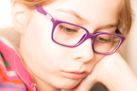 Face of eight years old, caucasian, blond and thoughtful child girl (kid) in glasses while reading a book. Close up portrait (macro). Childhood and education concept.の写真素材